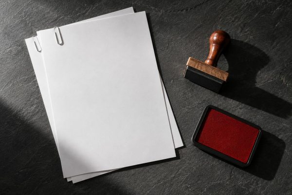 Hojas en blanco sujetas con clips junto a un sello de madera y un tampón rojo sobre pizarra